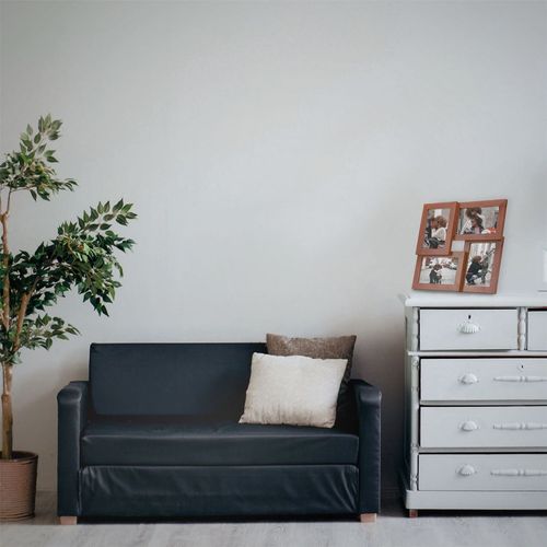 A minimalist living room has a dark sofa accented with white and brown pillows. A potted plant sits left, while a white dresser with wood veneer, topped with Maison & Whites Multi Picture Aperture 6 x 4 Photo Frame in wood, is on the right against a plain light wall.