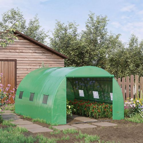An Outsunny 4x3x2m Green Tunnel Greenhouse, filled with flowers, rests beside a wooden shed amidst paving stones. Encircled by a wooden fence and trees under a partly cloudy sky, it provides ideal protection for enthusiastic gardeners.