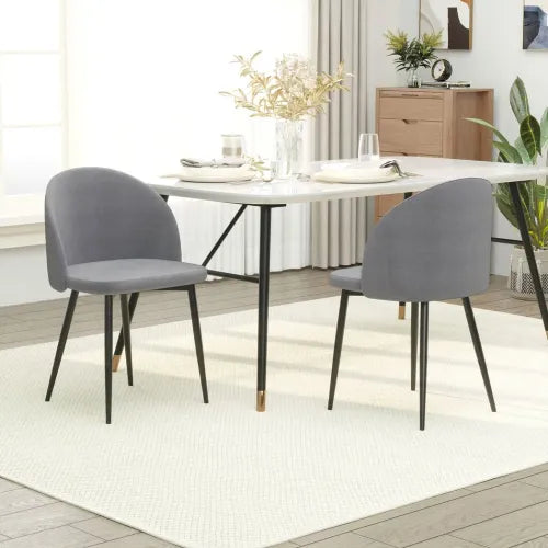 A modern dining room features a white steel frame table with gray armless 2 Pieces Modern Upholstered Fabric Bucket Seat Dining Chairs Living Room Grey. Golden accents highlight the chair legs. In the background, plants and a wooden sideboard are visible near a large window. White plates and glasses adorn the table.