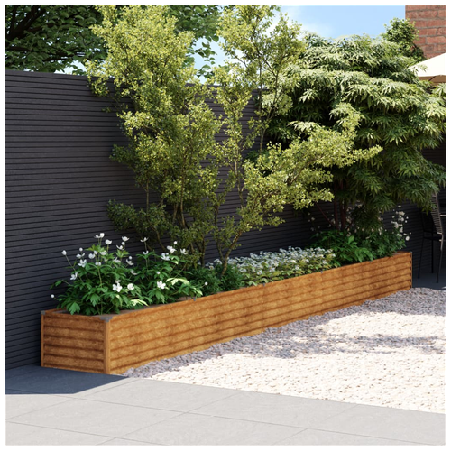 A sleek dark fence complements the vidaXL Garden Raised Bed 482x50x36 cm Corten Steel, filled with small trees and white flowers. Pebble landscaping and a patio umbrella enhance the modern outdoor setting.