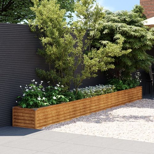 The vidaXL Garden Raised Bed 482x50x36 cm in Corten Steel, filled with small trees and white flowers, sits on a gravel-paved patio. A gray wooden fence reminiscent of weathered steel provides a backdrop, with lush green leaves and a clear blue sky overhead.